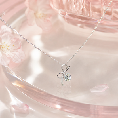桜の花びらとピンクのガラスの上に置かれた、輝くリボン結びデザインのモアサナイトネックレス。プレゼントに最適。